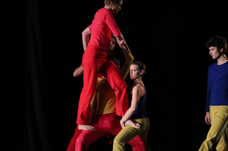 One dancer stands another's thighs as if she is she walking on air. One dark haired male dancer in blue and yellow observe